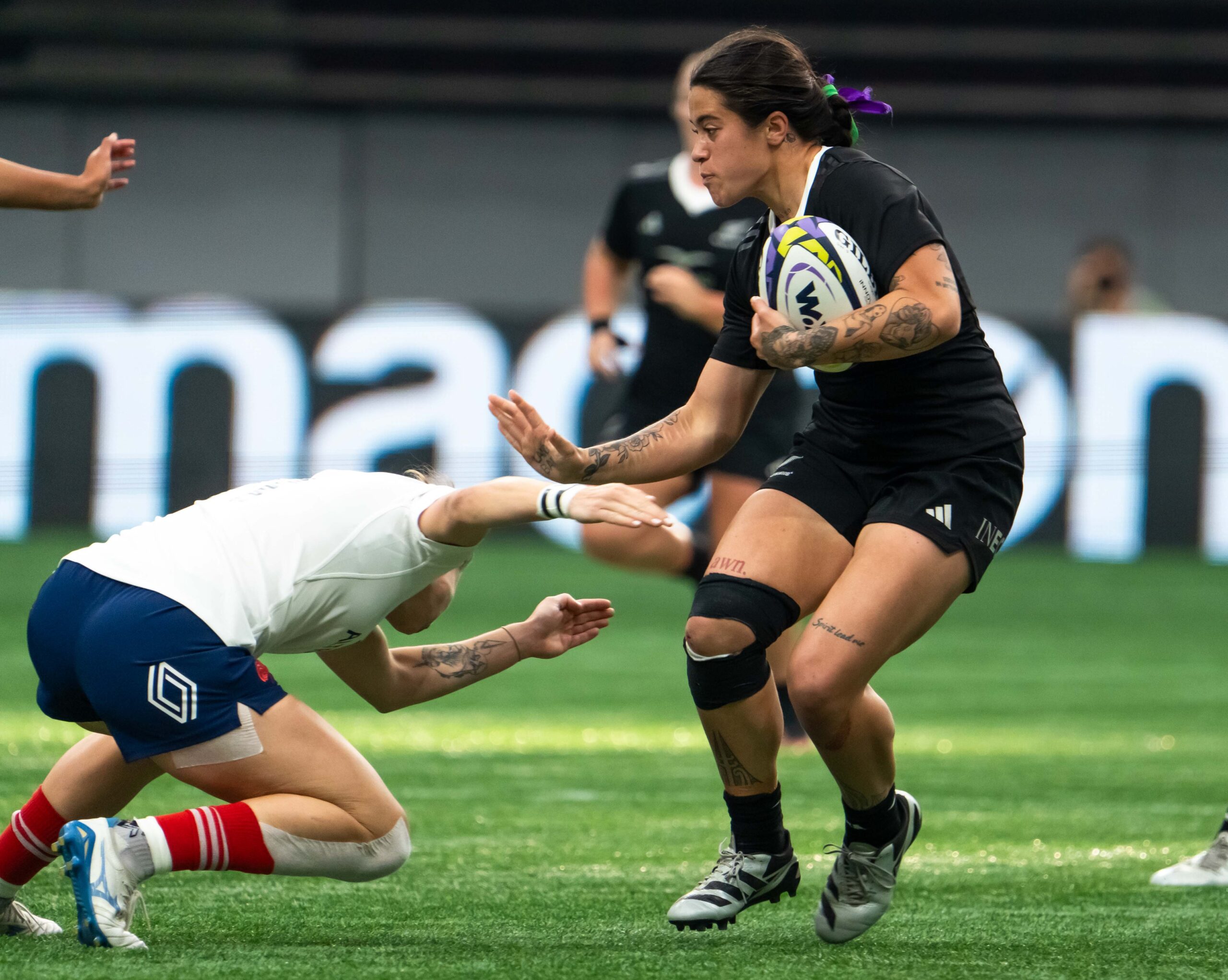 New Zealand Black Ferns player with tattoos fending off a tackle while carrying the Gilbert rugby ball.