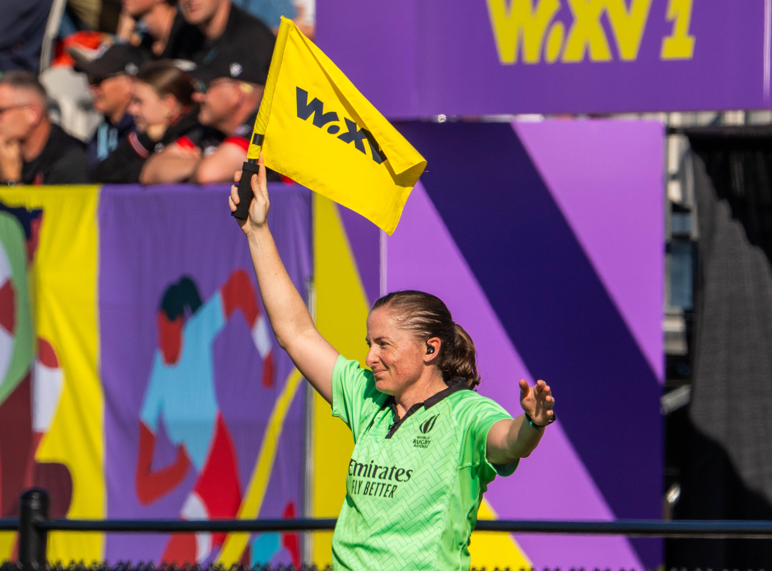 A female World Rugby assistant referee in a green Emirates jersey holding up a yellow WXV flag during a match in Dubai.