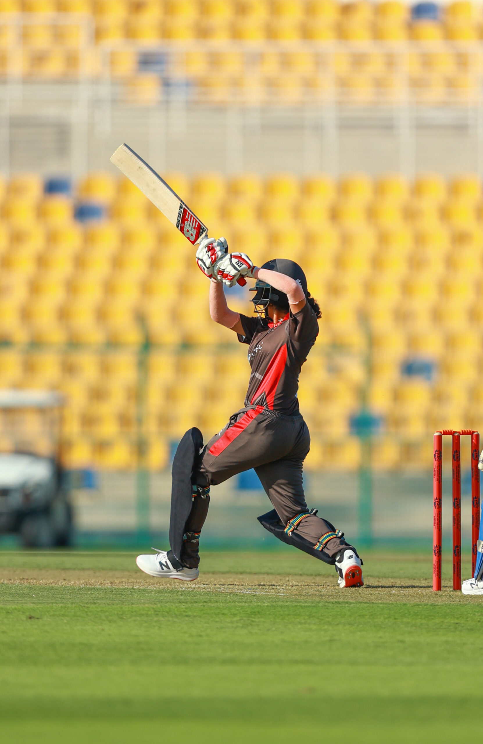A women's cricket batter playing a powerful pull shot against a yellow-seated stadium backdrop in Dubai.