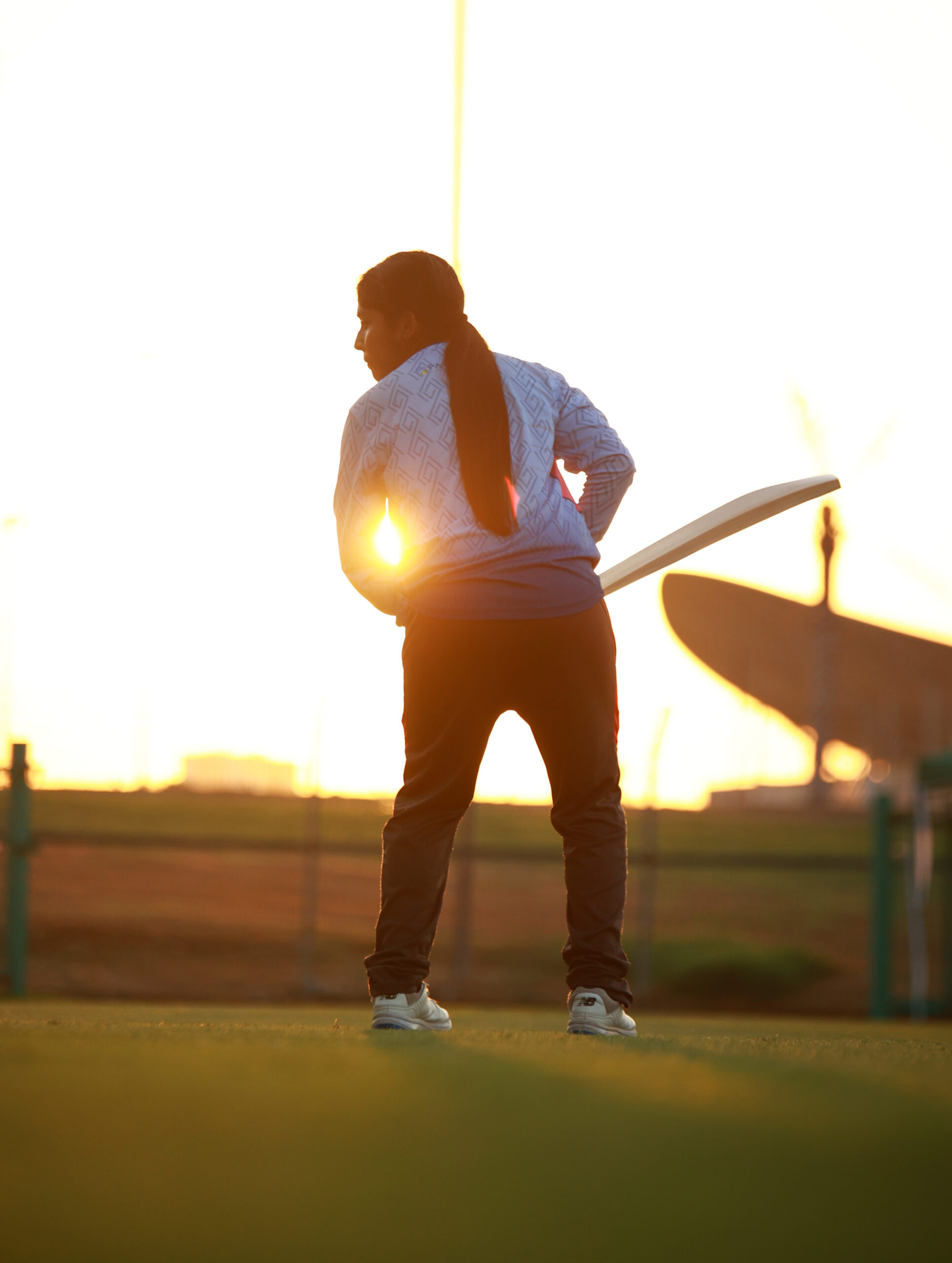 Silhouette of a female cricket player in batting stance against a Dubai desert sunset at the cricket ground.