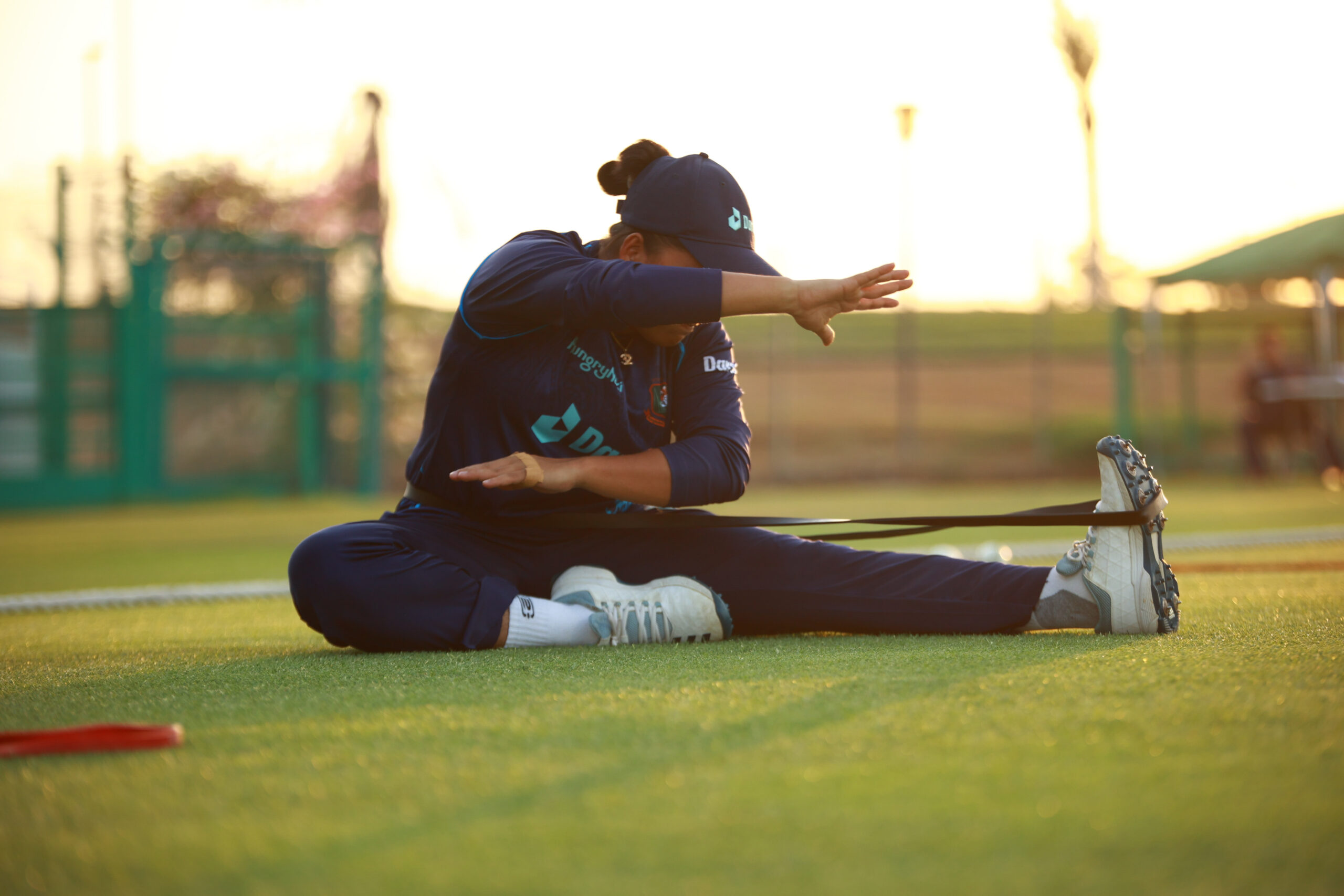 Professional cricketer in a blue kit performing a seated hamstring stretch with a resistance band at sunset in a Dubai sports complex
