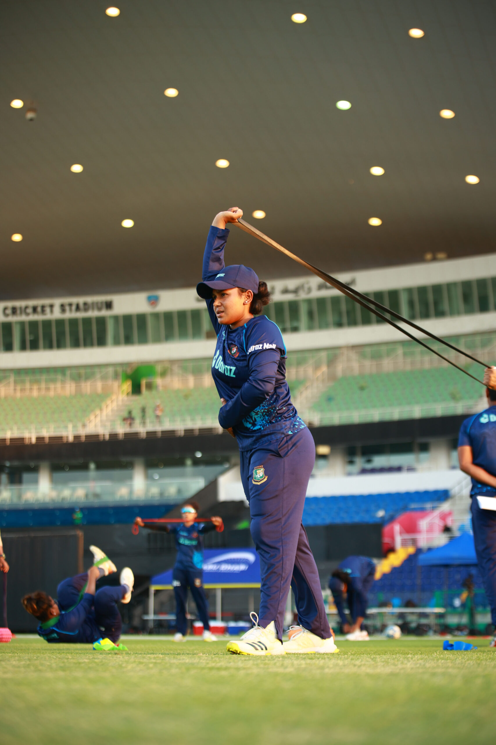 Professional female cricketer in a blue Bangladesh kit performing overhead mobility drills with a resistance band during warm-ups in a UAE stadium
