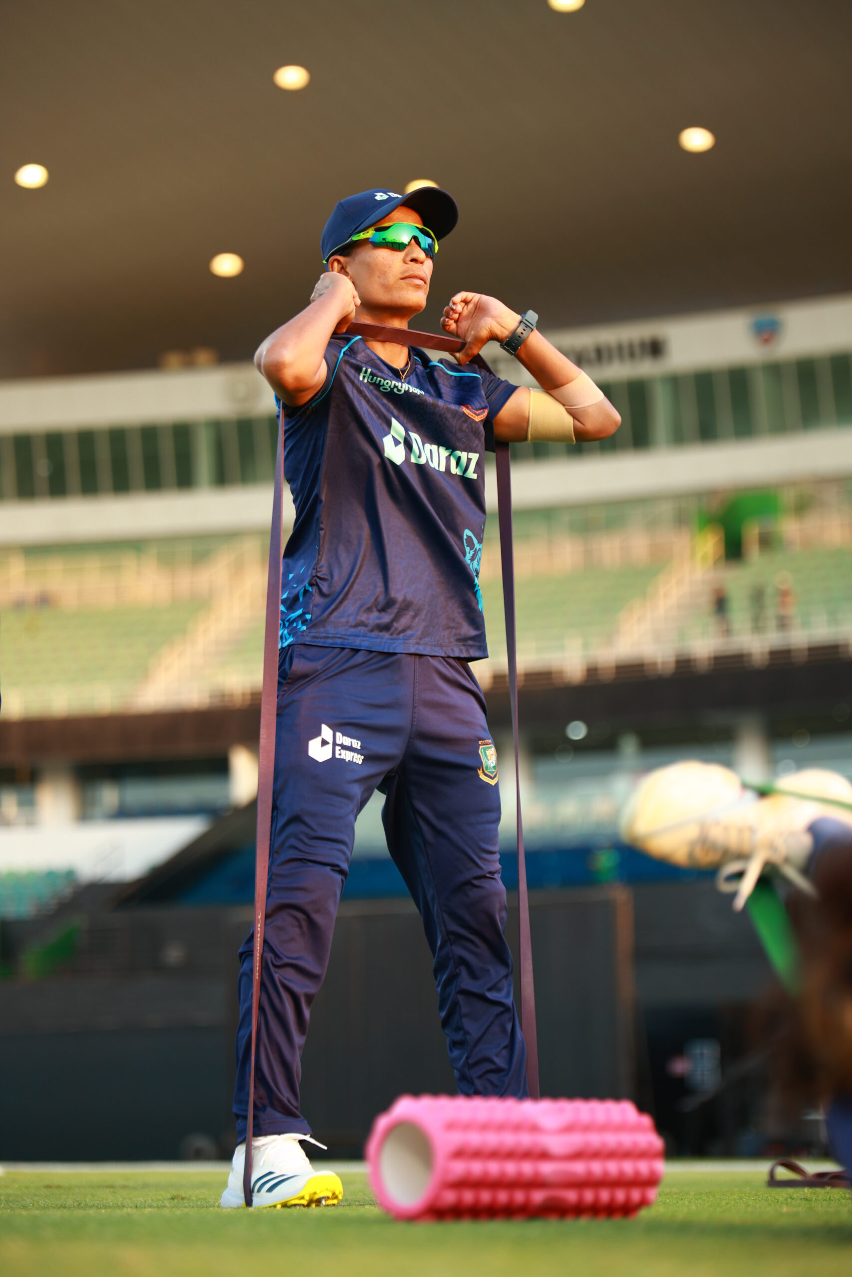 Professional female cricketer in a blue Bangladesh kit using a resistance band for strength training at a Dubai stadium