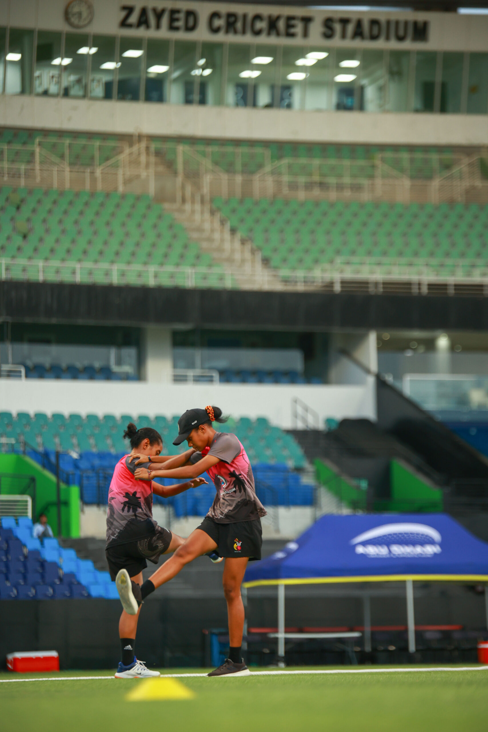 Two female cricket players in pink and grey kits performing leg stretches during a training session at a Dubai cricket stadium