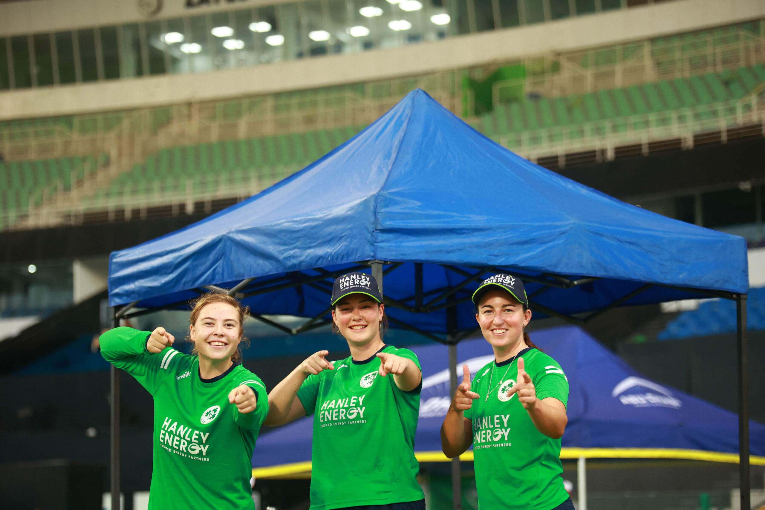 Three Ireland women's cricketers in green kits pointing at the camera and smiling under a team tent.