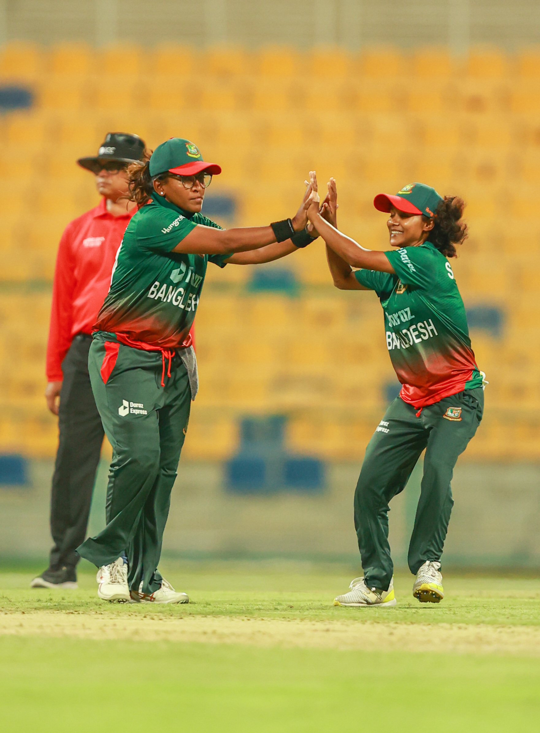 Two Bangladesh women cricketers celebrating a wicket with a high-five under stadium floodlights.