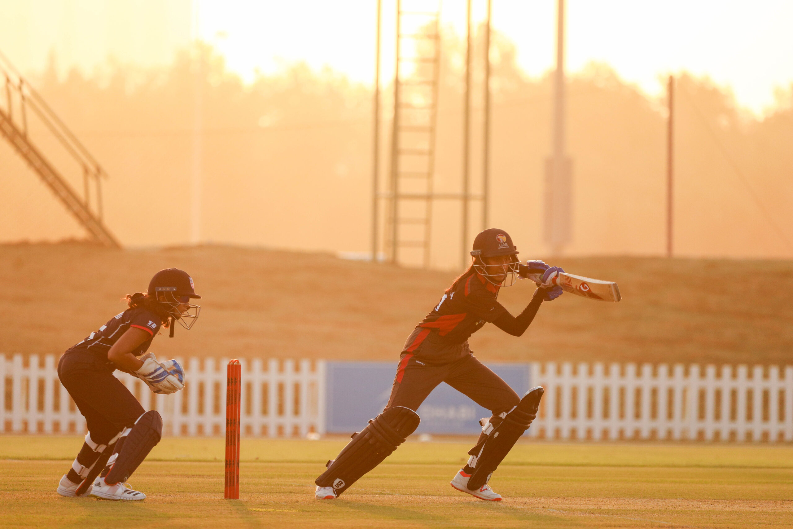 International women's cricket match at sunset in Dubai featuring UAE and USA national team players.